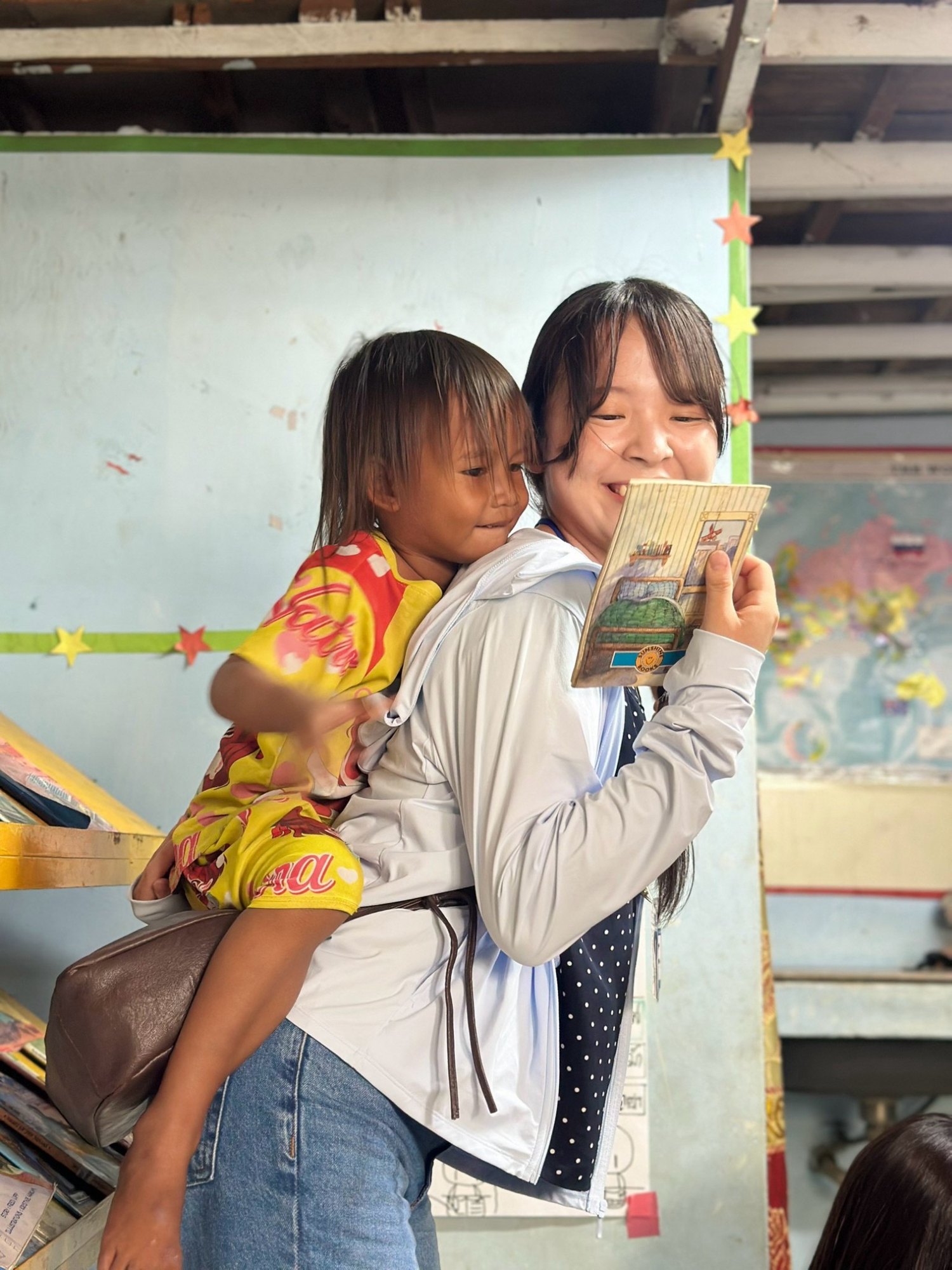 女子学生が現地の子どもをおんぶしながら、いっしょに絵本を読んでいる場面